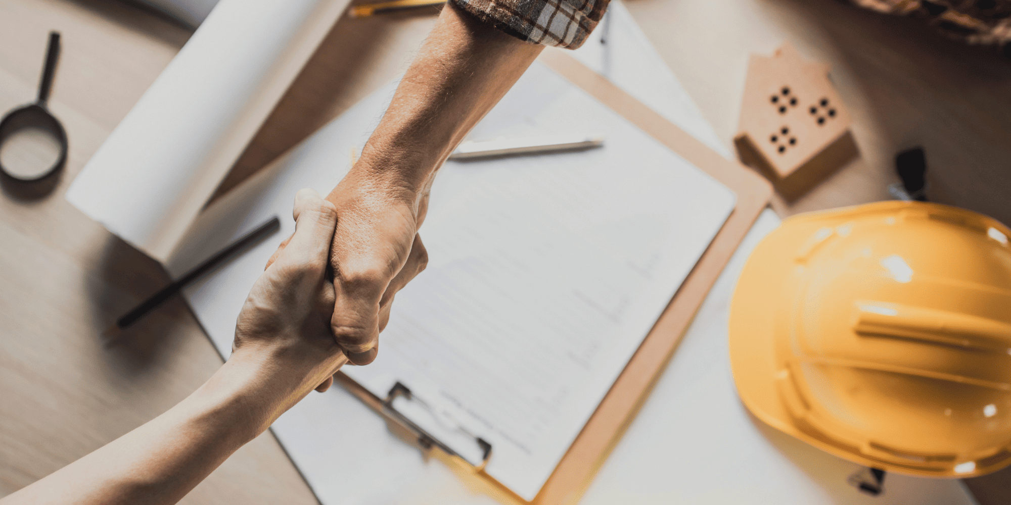 Stock Image of Two People Shaking Hands Over a Table with Documents and a Hard Hat